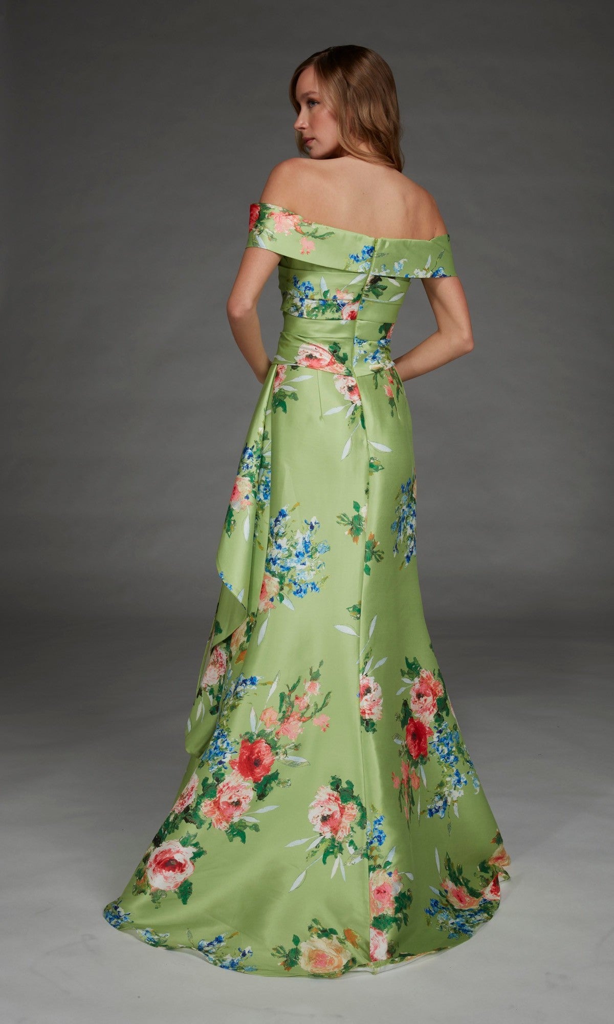 Wearing the Alyce 27915 long formal dress, a woman stands facing away against a plain gray background. The floor-length satin gown features an off-the-shoulder design with a green floral print.